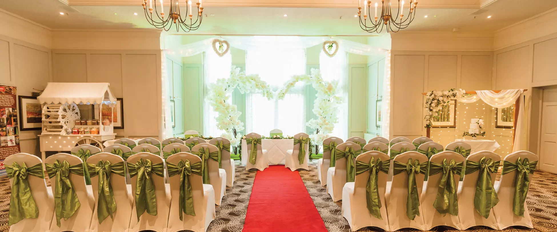 A room set up for a wedding. Chairs line either side of a red carpet. The chairs are covered in white fabric and have green bows. The carpet leads to a central table framed by a large flower heart-shaped arch.