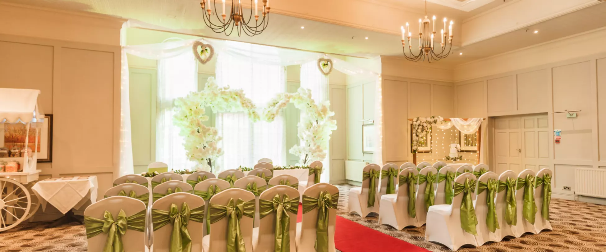 A large room set up for a wedding ceremony. At the front of the room is a heart shaped arch of white flowers. There are dozens of chairs covered in white cloth and tied with green ribbons.
